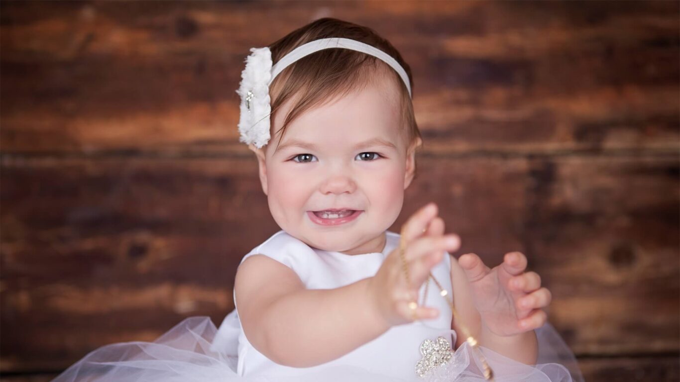 Smiling toddler in a white dress and headband reaching forward, with a wooden background.