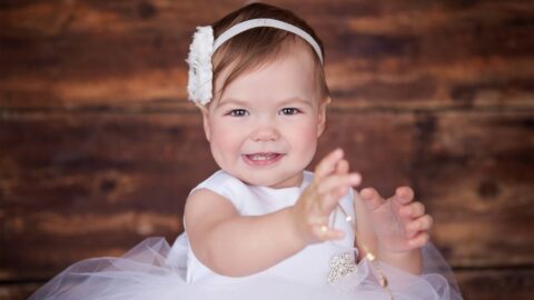 Smiling toddler in a white dress and headband reaching forward, with a wooden background.