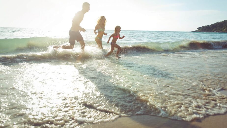 Family joyfully running on sandy beach, splashing in gentle waves under bright sunshine; perfect coastal holiday experience.