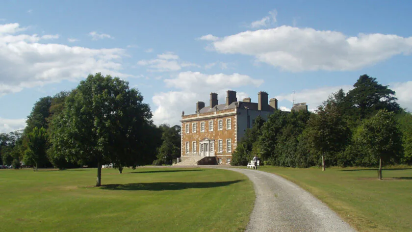 Elegant country house hotel with tree-lined drive, lush gardens, and peaceful surroundings on a sunny day.