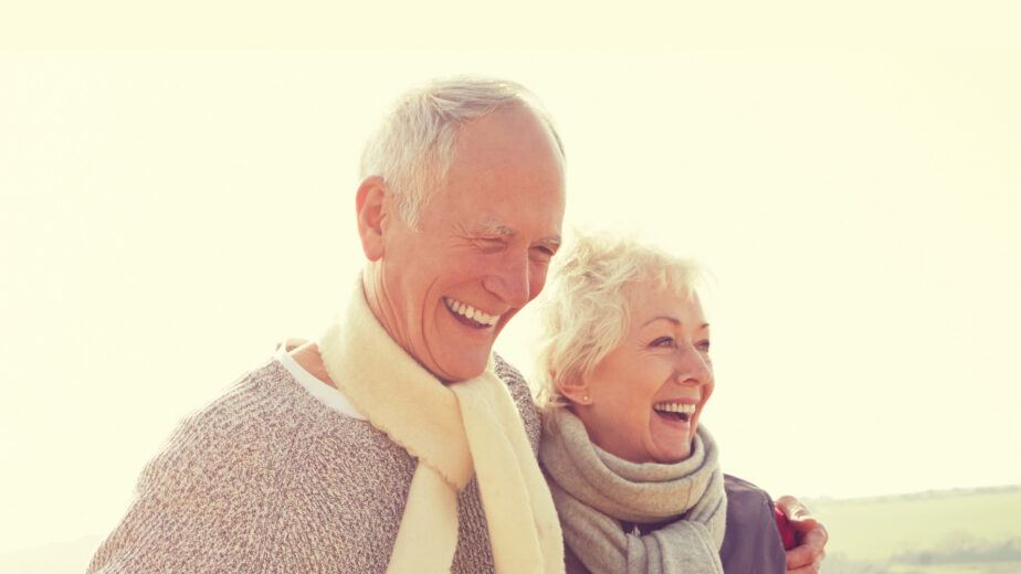 An older man and woman stand outdoors, smiling and wearing scarves and sweaters, with a light sky and green landscape in the background.