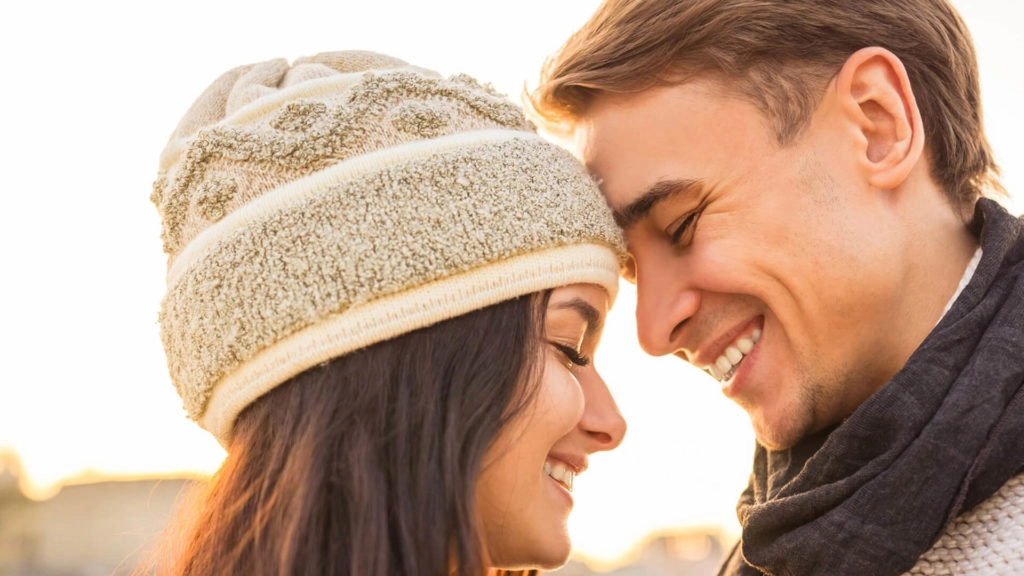 A close-up of a smiling couple touching foreheads outdoors, both wearing winter clothing including a hat and scarf.