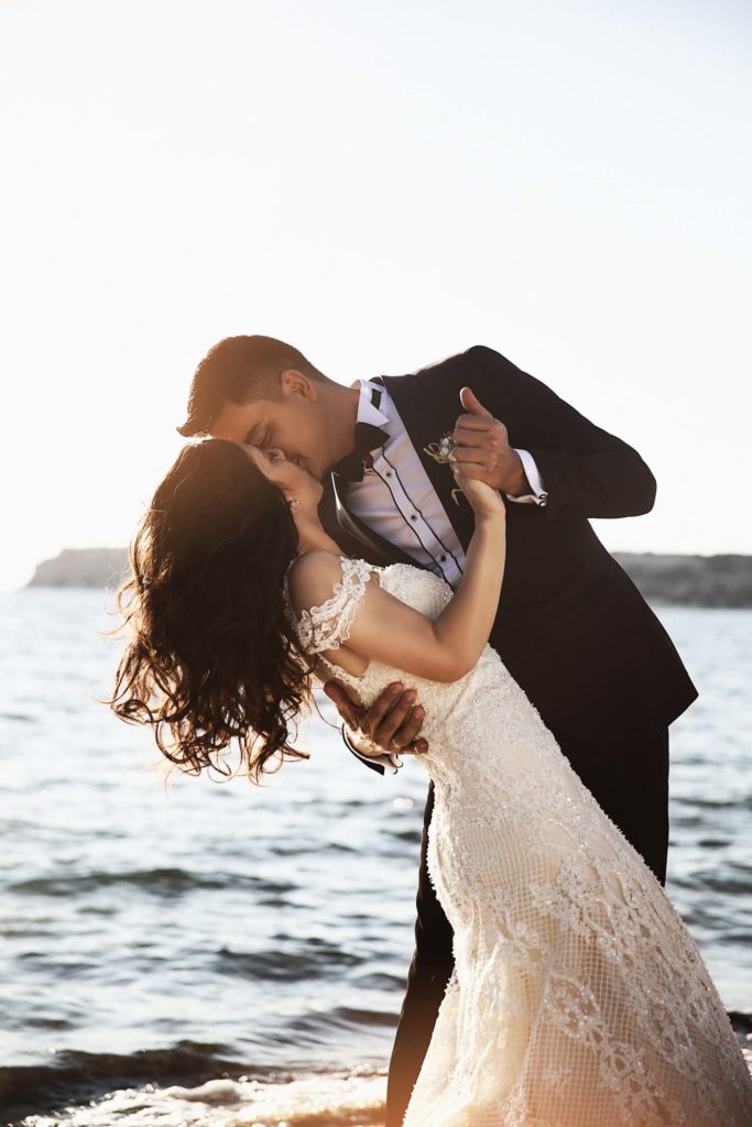 A couple in formal wedding attire shares a kiss while dancing on a beach with the ocean and bright sunlight in the background.