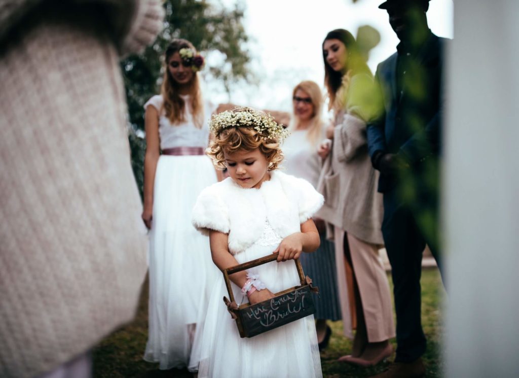 A young flower girl in a white dress and floral crown holds a small basket, while several adults and another girl stand in the background outdoors.