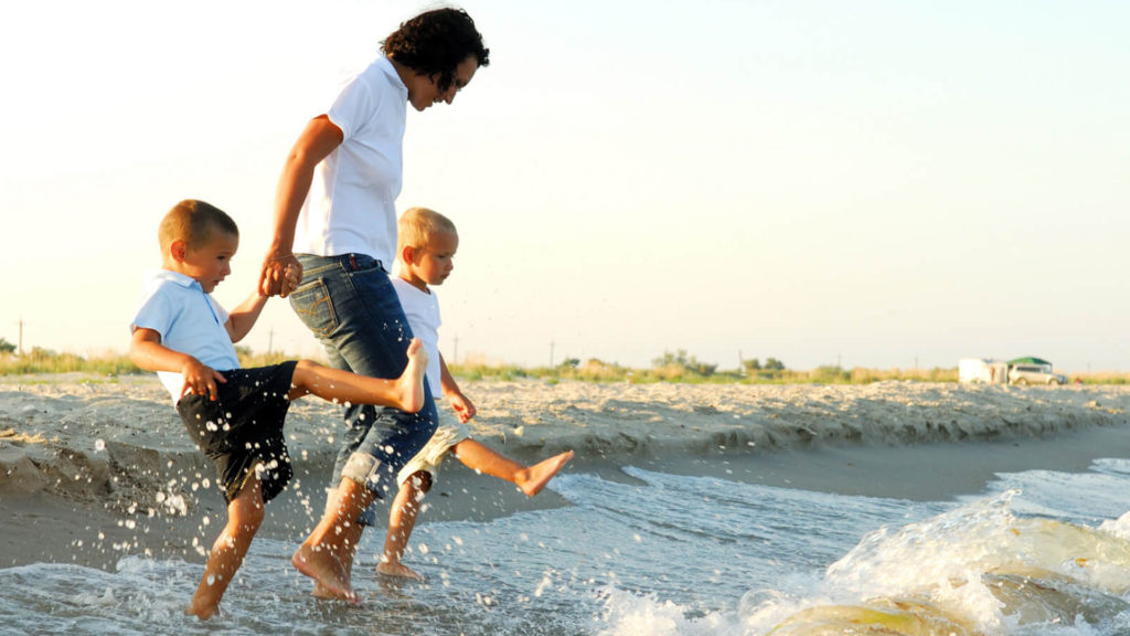 autumn-family An adult and two children wearing white shirts and jeans walk along the shore, kicking water as waves reach their feet on a sandy beach.