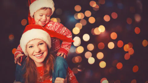 Woman wearing a Santa hat gives a piggyback ride to a young child, also in a Santa hat, with festive lights blurred in the background.