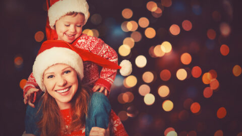 Woman wearing a Santa hat gives a piggyback ride to a young child, also in a Santa hat, with festive lights blurred in the background.