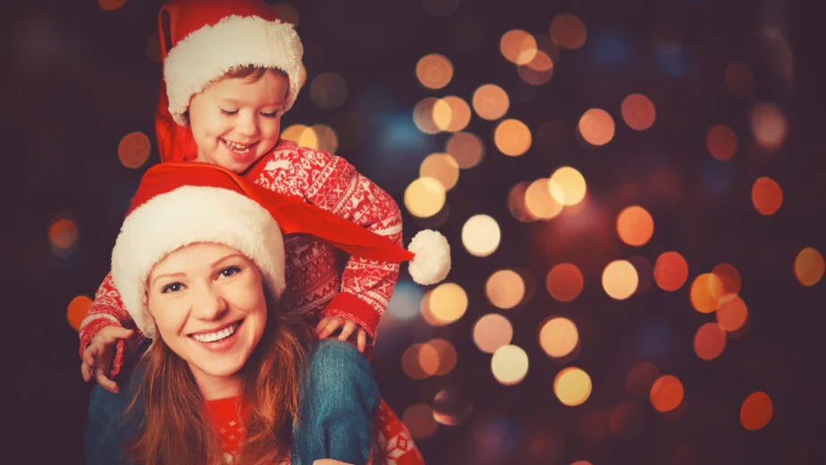 Woman wearing a Santa hat gives a piggyback ride to a young child, also in a Santa hat, with festive lights blurred in the background.
