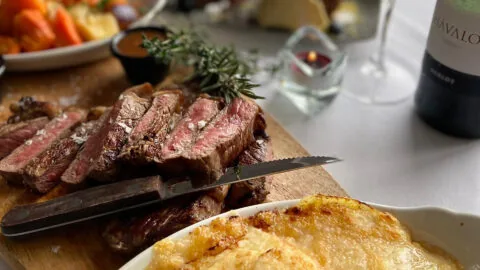 Sliced medium-rare steak on a wooden board with a knife, accompanied by a dish of gratin potatoes and a bottle of wine on a set dining table.