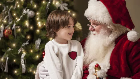 A young boy sits on Santa Claus’s lap, smiling, in front of a decorated Christmas tree with lights and ornaments.