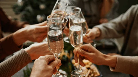 A group of people clink champagne glasses in a toast, with food and a decorated tree visible in the background.