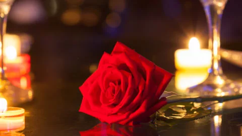 A red rose lies on a reflective table surrounded by lit candles and champagne glasses in a dimly lit setting.