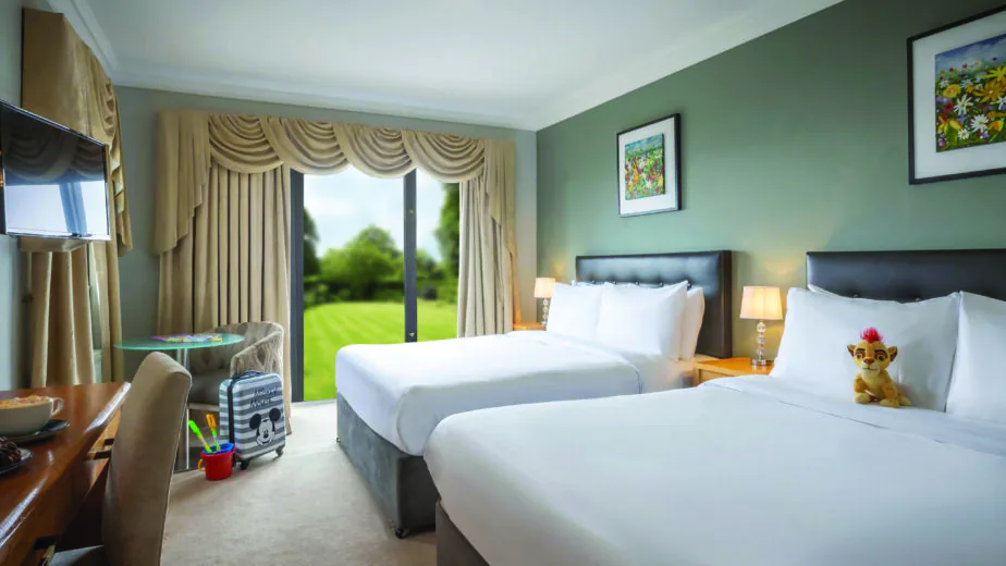Bright hotel room with two cosy beds, garden view, family-friendly decor, flat-screen TV, and modern furnishings.