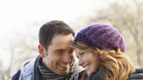 A man and woman sit close together on a bench outdoors, dressed in winter clothing, smiling and looking at each other.