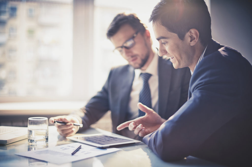 Things to Do  Two men in business suits sit at a desk, discussing documents and using a digital tablet in a bright office setting.