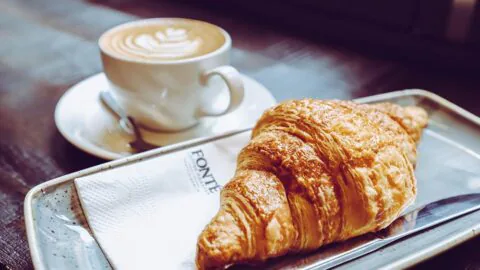 A cup of coffee with latte art sits on a saucer next to a croissant on a metal tray with a Fonté brandednnapkin and knife.