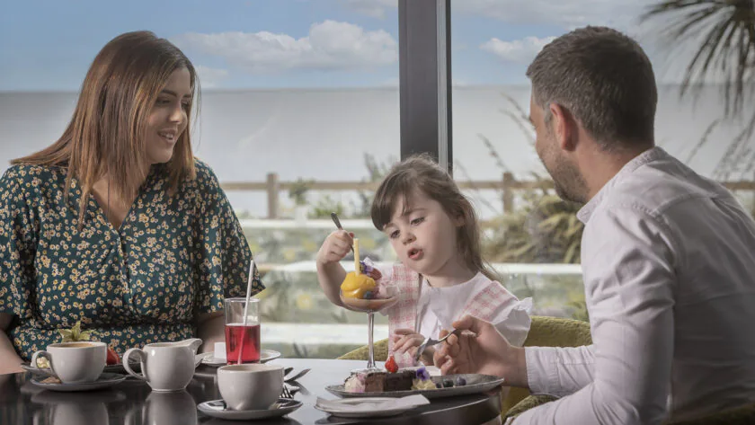 Family enjoys tea with sea view; smiling parents and child relish desserts in stylish hotel dining, coastal escape.