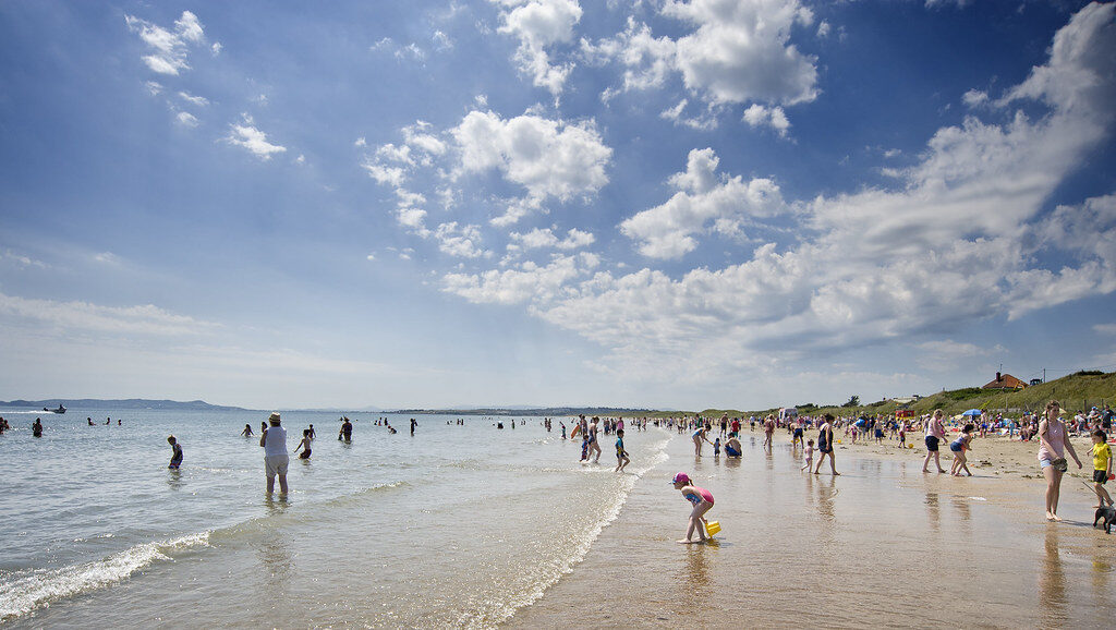 Guests enjoy a sunny day at a busy beach, perfect for swimming and sandcastle building, with clear skies and gentle waves.