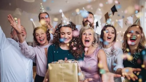 A group of people wearing party hats celebrate indoors, smiling and throwing confetti, with one person holding a wrapped gift box.