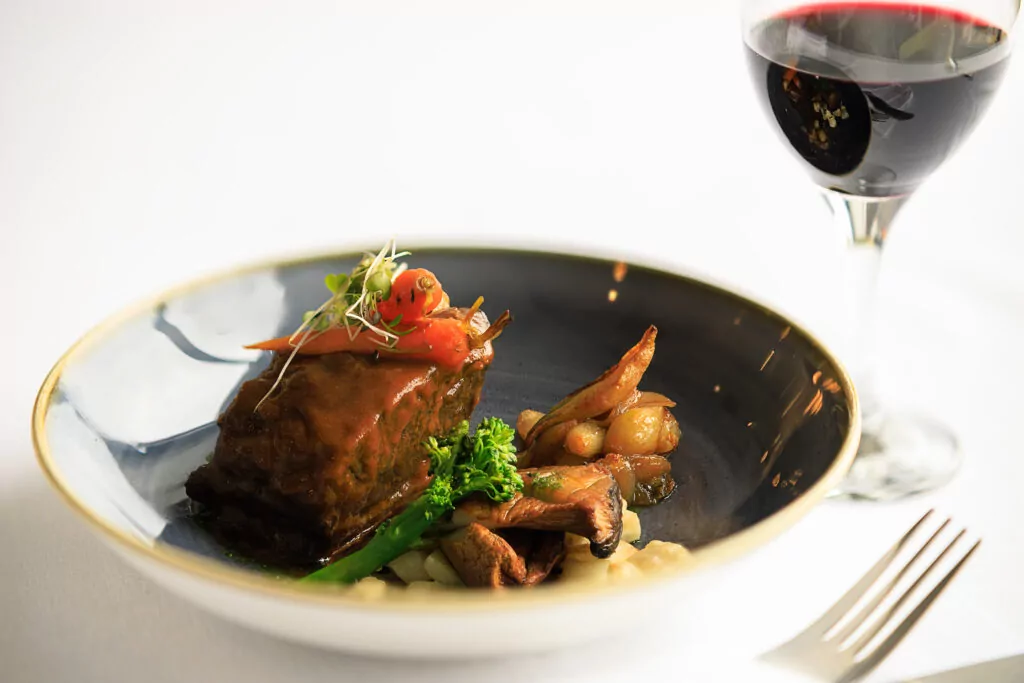 A plate with braised meat, vegetables, and sauce is next to a glass of red wine on a white tablecloth.