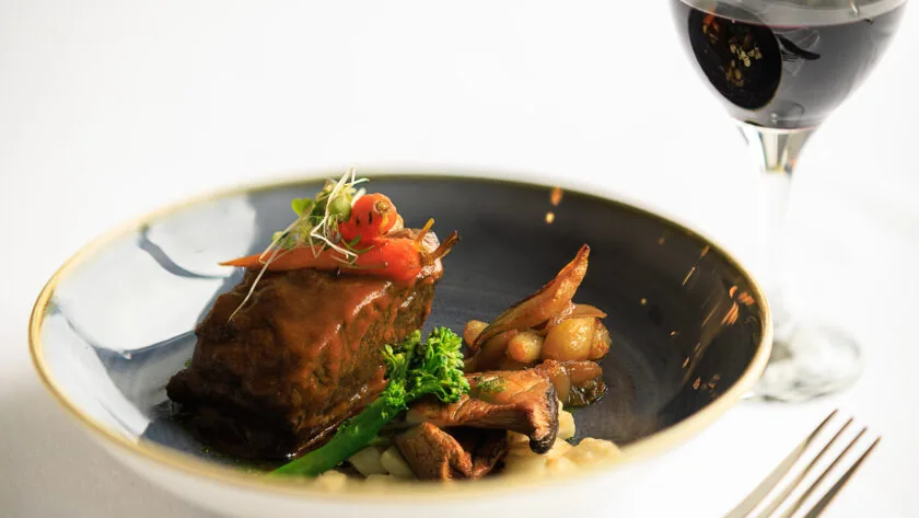 A plate with braised meat, vegetables, and sauce is next to a glass of red wine on a white tablecloth.