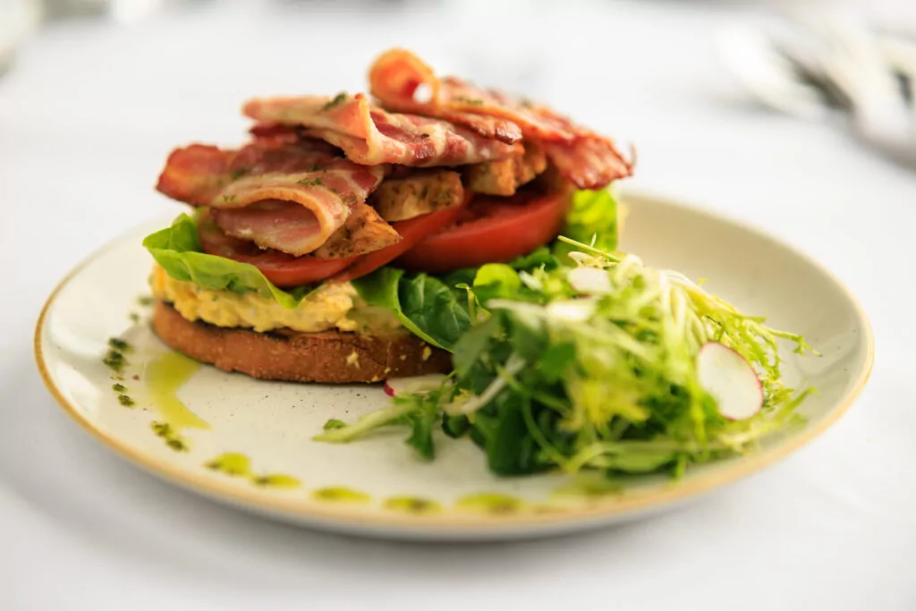 Brown bread with scrambled eggs, tomatoes, bacon and a side salad on a plate on a white table cloth