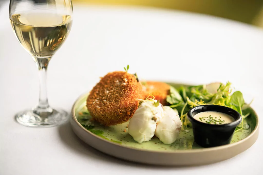 A plate of breaded fried cakes, poached eggs, green salad, a small cup of sauce, and a glass of white wine on a white tablecloth.
