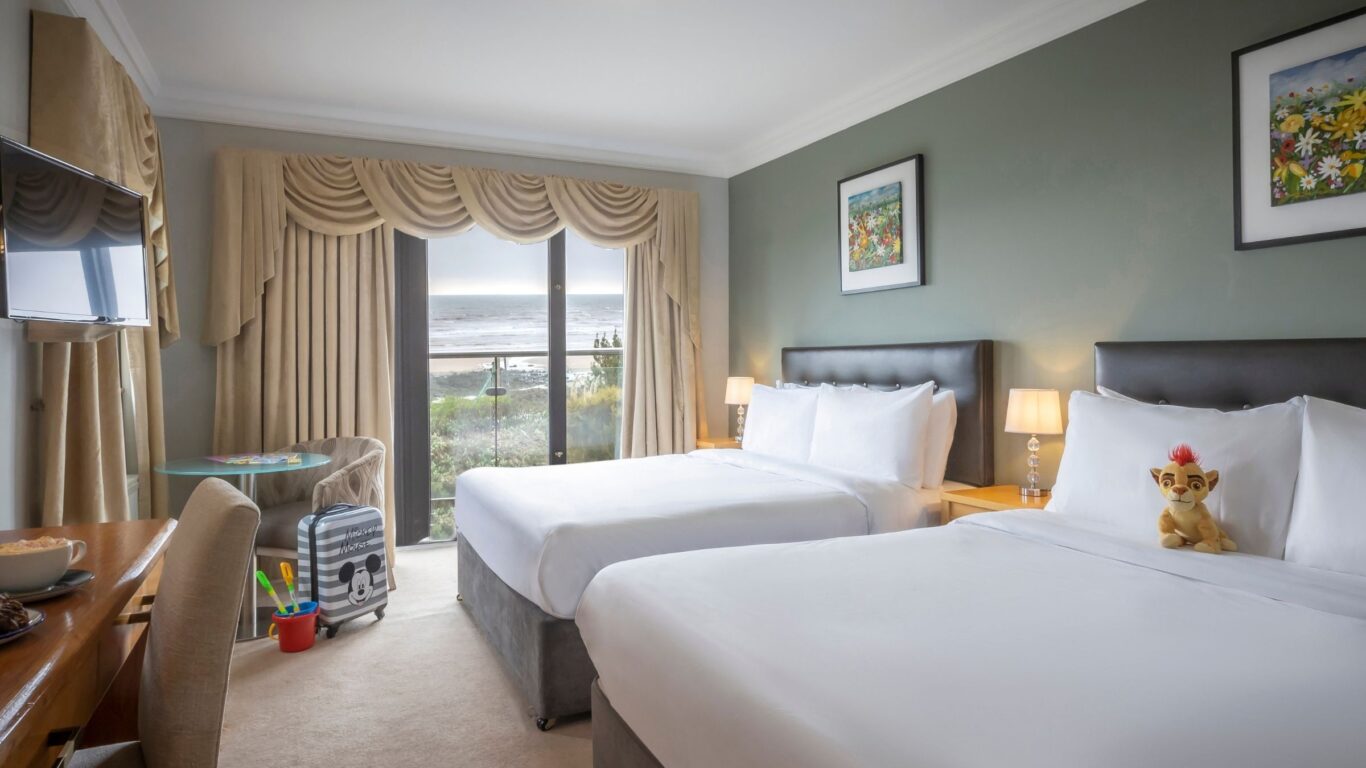 Spacious hotel room with two double beds, sea view balcony, elegant decor, cosy seating, and vibrant local artwork.