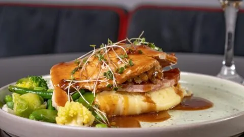 A plated meal featuring sliced roast meat on mashed potatoes with gravy, garnished with microgreens, served with mixed vegetables and a glass of white wine in the background.