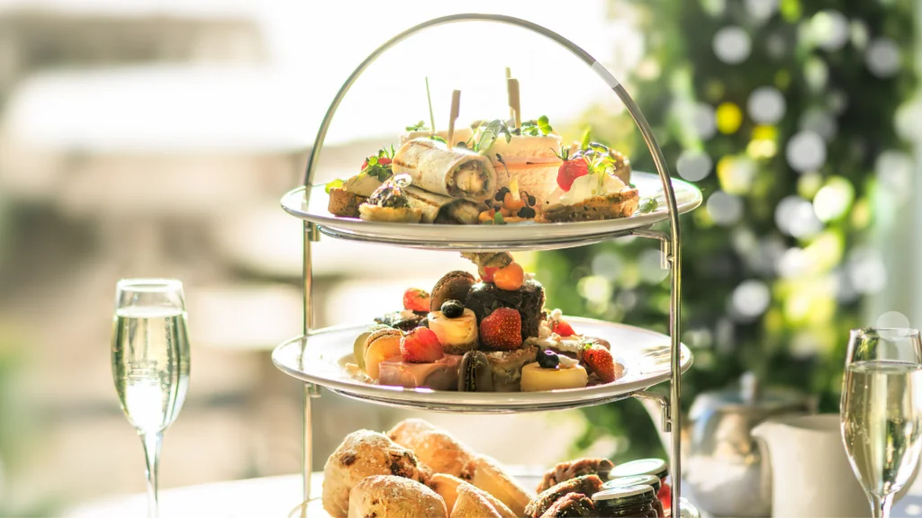 A three-tiered stand with assorted finger sandwiches, pastries, fruits, and scones, accompanied by two glasses of sparkling drink on a table.