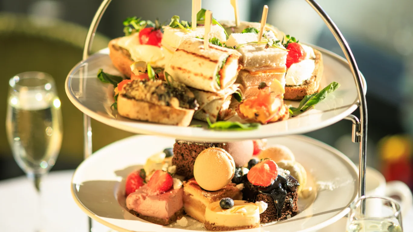 Two-tiered tray with assorted finger sandwiches, pastries, macarons, and fruit, set on a table with a glass of sparkling beverage in the background.