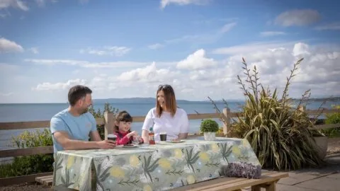 Family enjoying seaside dining at a picnic table with stunning ocean views and blue skies, perfect for holiday relaxation.