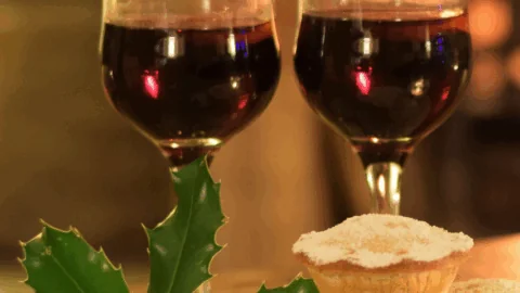 Two glasses of red wine and three small mince pies dusted with powdered sugar, with a sprig of holly placed beside them on a table.