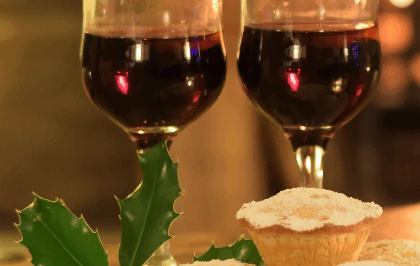 Two glasses of red wine and three small mince pies dusted with powdered sugar, with a sprig of holly placed beside them on a table.