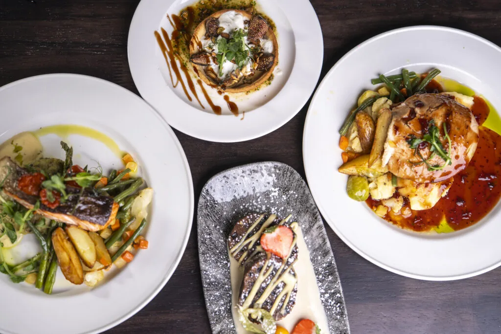 Four plates of gourmet food sit on a dark wooden table, featuring entrees of fish, poultry, and mushrooms, along with a dessert garnished with a strawberry.