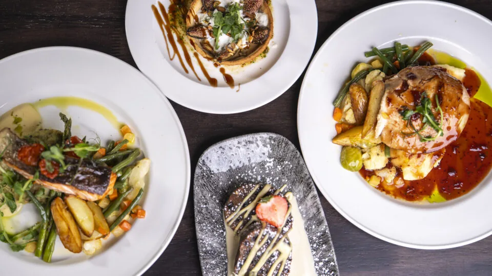 Four plates of gourmet food sit on a dark wooden table, featuring entrees of fish, poultry, and mushrooms, along with a dessert garnished with a strawberry.