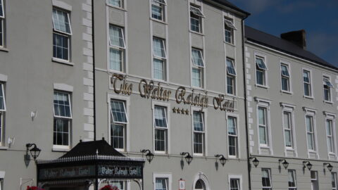 The exterior of The Walter Raleigh Hotel, a multi-story grey building with white trim, large windows, and a sign above the entrance.