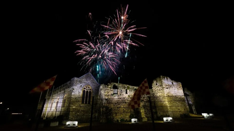 Fireworks light up the night sky above an illuminated stone ruin, with checkered flags visible in the foreground.