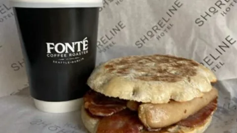 A bacon and sausage breakfast sandwich on an English muffin next to a black takeaway coffee cup labeled 