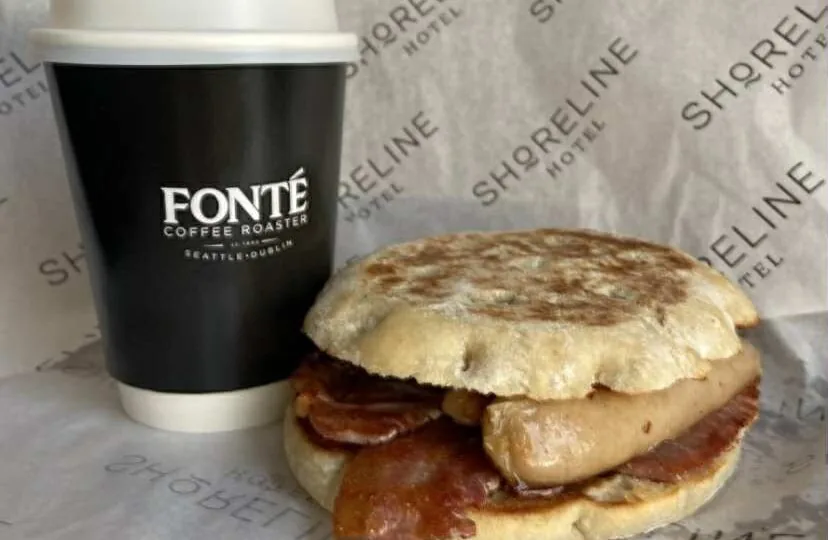A bacon and sausage breakfast sandwich on an English muffin next to a black takeaway coffee cup labeled