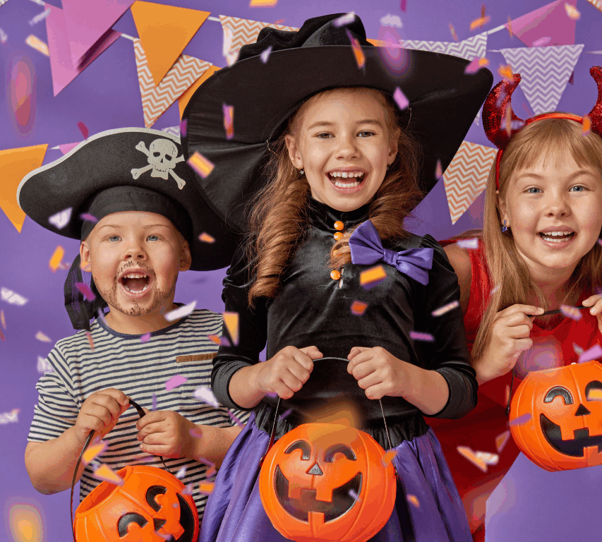 Three children in Halloween costumes—a pirate, a witch, and a devil—holding pumpkin buckets, smiling, with confetti and festive flags in the background.
