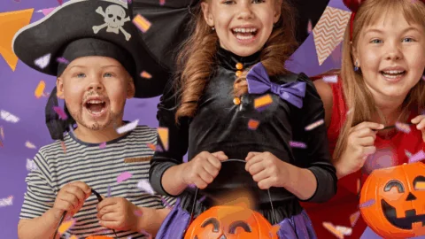 Three children in Halloween costumes—a pirate, a witch, and a devil—holding pumpkin buckets, smiling, with confetti and festive flags in the background.