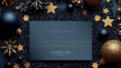 A Shoreline Hotel gift card rests on black glitter with gold ornaments, stars, pinecones, and baubles arranged decoratively around it.
