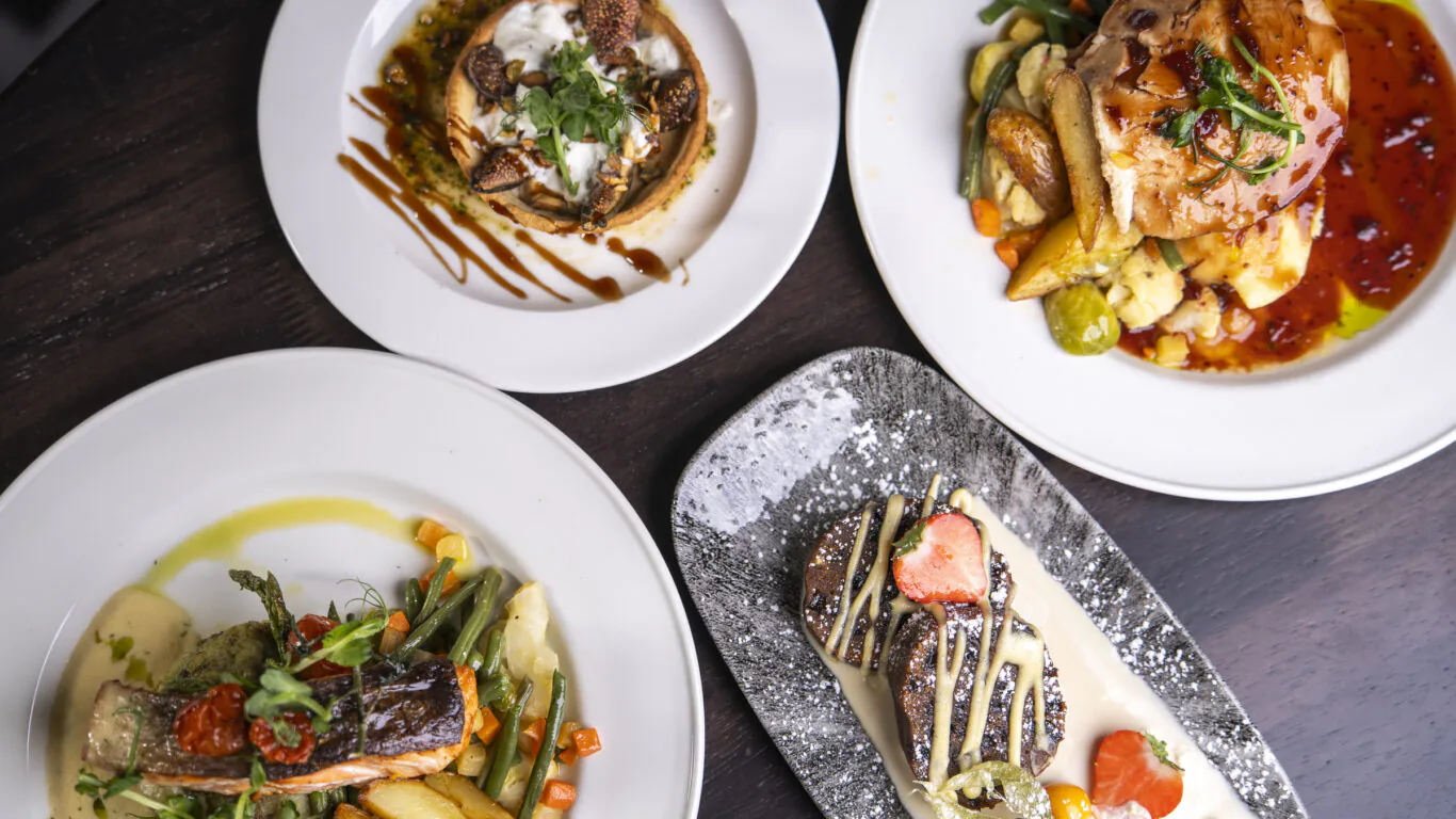 Four plated gourmet dishes, including a dessert with fruit garnish and whipped cream, arranged on a dark wooden table.