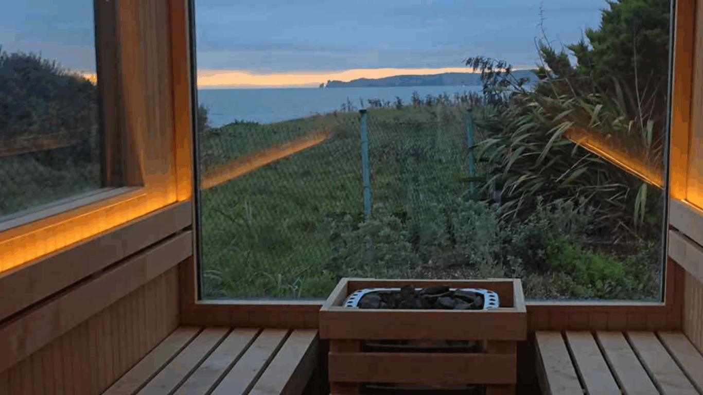 A wooden sauna interior with a central heater and large windows offering a view of greenery, a fence, and the sea under a cloudy sky at sunset.