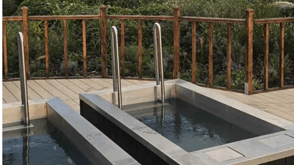 Two rectangular outdoor plunge pools with metal handrails are set on a wooden deck, bordered by a wooden fence and surrounded by greenery.