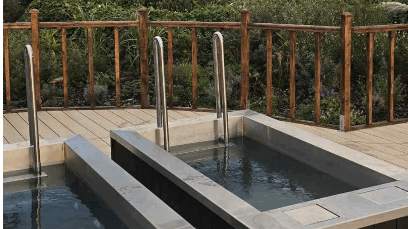 Two rectangular outdoor plunge pools with metal handrails are set on a wooden deck, bordered by a wooden fence and surrounded by greenery.