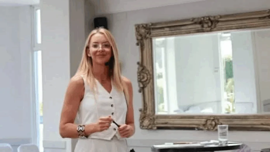 A woman in a white sleeveless jumpsuit stands beside a round table with a glass of water and microphone in a bright, carpeted room.