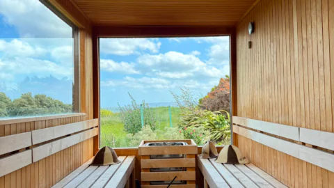 Relaxing wooden sauna with window views of lush green landscape and blue skies at luxurious hotel retreat.