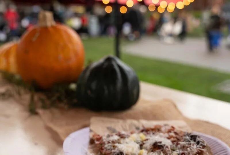Fresh pizza slice with cheese on a wooden table, surrounded by pumpkins, and vibrant festival tents in a garden setting.